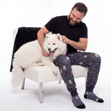 Man sitting on a chair with a white dog, wearing pajama pants and socks with batman pattern and white dog pattern.