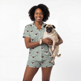 Woman wearing a pug-themed hort button down pajama set holding a pug on a white background