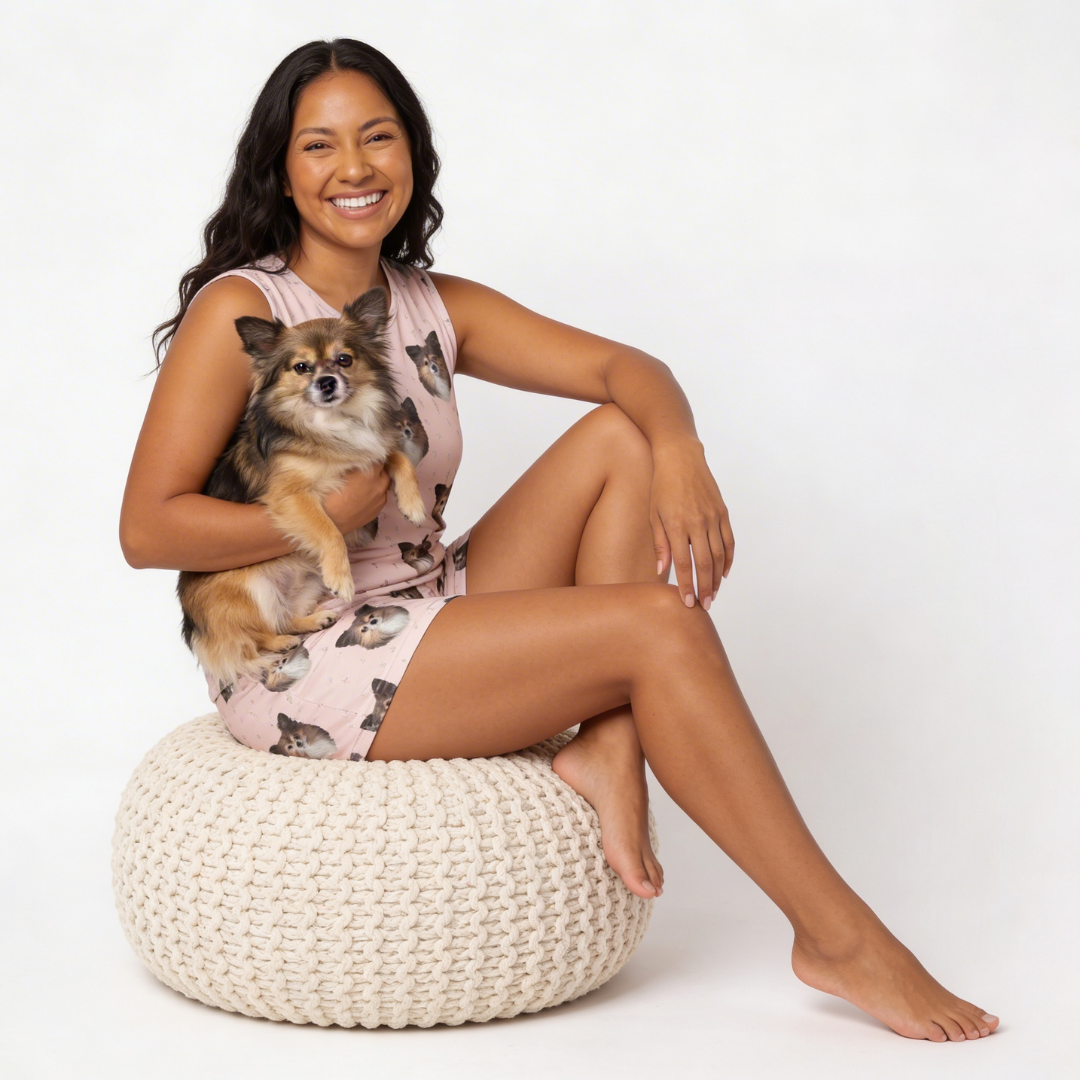 Woman wearing tank top pajamas sitting on a woven ottoman holding a small dog, wearing a pink dress with dog print.