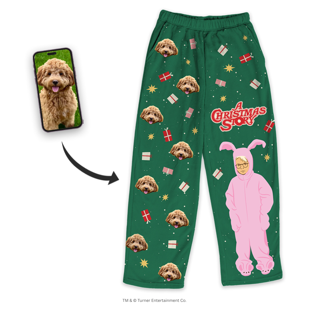 Green pajama pants featuring a repeating dog's face pattern, gift boxes, snowflakes and the text 'A Christmas Story' with a character in a pink bunny costume, shown next to a smartphone displaying the dog's photo.