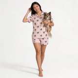 Woman wearing pink short button down pajama set with dog pattern, holding a small dog, on a white background