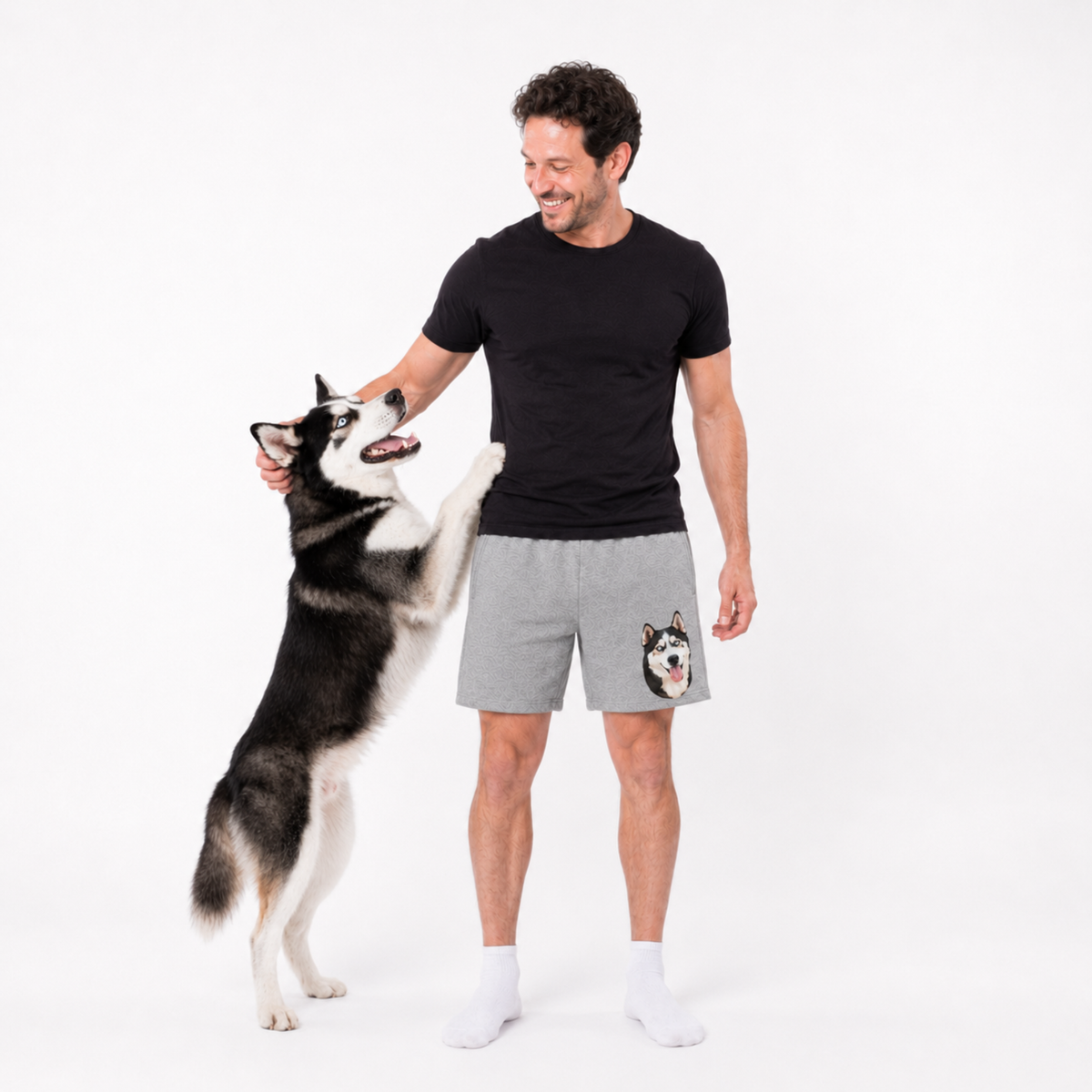 Man wearing a black t-shirt and gray shorts with a dog graphic, standing next to a dog on a white background