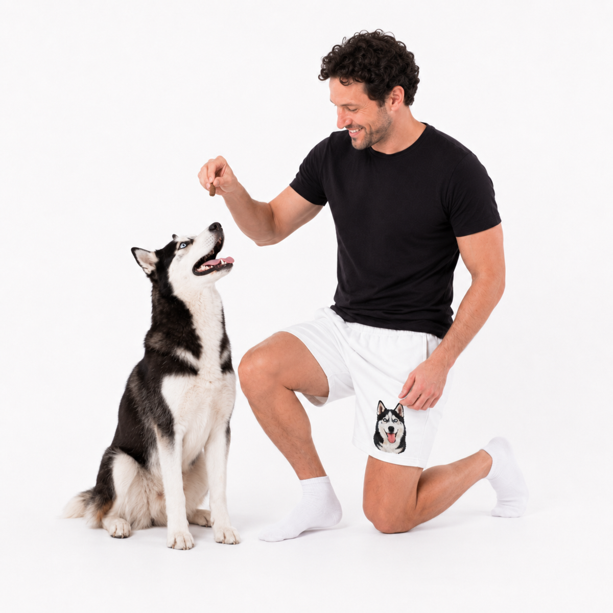Man sitting on the floor wearing a black t-shirt and white shorts giving a treat to a dog.