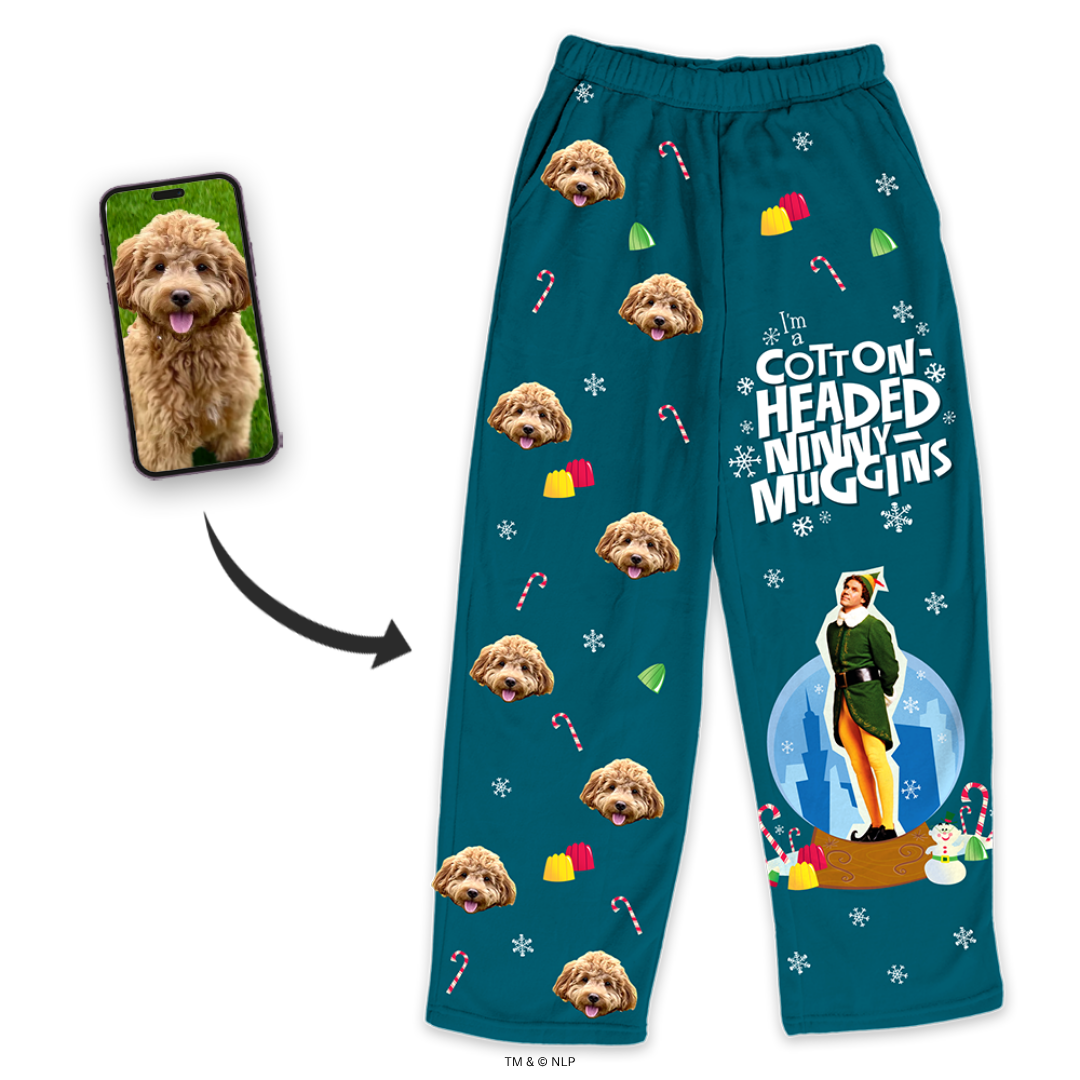 Teal pajama pants featuring a dog's face pattern and the phrase 'Cotton Headed Ninny Muggins' with a character from a Christmas movie at the bottom, shown beside a smartphone displaying the dog's photo.