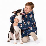 A smiling young boy in a navy blue Christmas dog pajama hoodie hugs a black and white dog on a clean white background. The hoodie features a pattern of dogs wearing Santa hats, with 'Merry Christmas' text visible on the sleeve.