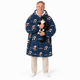 A smiling man wearing a navy blue Christmas-themed hooded robe with a penguin and Santa pattern, holding a black and white cat. He is wearing beige pants and white socks against a plain white background.