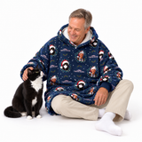 A man wearing a navy blue Christmas-themed hooded sweatshirt sits on a white studio floor next to a black and white cat. The sweatshirt features a festive print of penguins in Santa hats, snow globes, and holiday decorations.