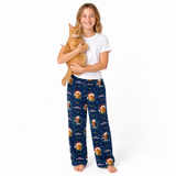 A young girl stands barefoot against a white studio background, holding an orange tabby cat. She is wearing a white short-sleeve T-shirt and navy blue pajama pants covered in a festive gingerbread man and Santa pattern.