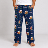 A person wearing navy blue Christmas-themed pajama pants with a repeating print of a golden dog in a Santa hat, green wreaths, holly leaves, and snowflakes. The person is standing barefoot against a light gray studio background.