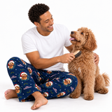 A man wearing blue Christmas-themed dog pajamas sits on the floor, smiling as he brushes a fluffy golden-brown dog with a brush against a white background.