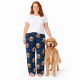 A woman stands barefoot next to a golden retriever dog wearing navy blue pajama pants with a repeating pattern of golden retriever faces in Santa hats, holly leaves, and snowflakes. The woman is also wearing a plain white t-shirt.