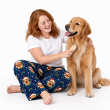 A woman with red hair sits cross-legged on a white studio floor, brushing a golden retriever dog. She wears a white T-shirt and navy pajama pants printed with a repeating pattern of golden retrievers wearing Santa hats and small holiday motifs.