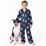 A young boy stands barefoot next to a black, white, and tan dog, both wearing matching navy blue pajamas with a festive print of dogs in Santa hats and snowflakes, against a solid white background.