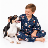 A young boy wearing a navy blue Christmas pajama set with a dog print sits cross-legged on a white studio floor, smiling at a black, white, and tan dog lying beside him.