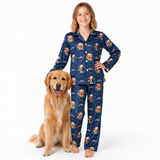 A young girl stands barefoot next to a golden retriever dog, wearing a navy blue long-sleeve pajama set with a festive Santa Claus dog print. The pajamas feature a dark blue background decorated with cartoon dogs in Santa hats, snowflakes, and stars.