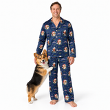 A man wearing a matching navy blue pajama set with a Santa Claus dog print stands barefoot next to a corgi on a plain white studio background.