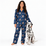 A woman wearing a navy blue pajama set with a Christmas dog print stands barefoot next to a Dalmatian dog on a white studio background. The pajamas feature a festive pattern of dogs in Santa hats, snowflakes, and stars.