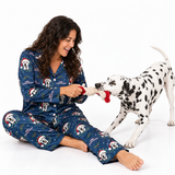 A woman wearing a navy blue Christmas-themed pajama set sits on the floor playing with a Dalmatian dog, which is holding a red and white tug toy. The pajamas feature a print of Dalmatians wearing Santa hats with 'Merry Christmas' text.