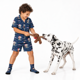 A child wearing a navy blue Christmas-themed pajama set with a dog print, playing tug-of-war with a brown plush dog toy on a white studio background.
