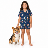 A young girl stands barefoot on a white studio background next to a corgi dog, wearing a matching navy blue pajama set with a Christmas dog print. The pajamas feature a short-sleeve button-up top and shorts with a repeating pattern of Santa Claus dogs, snowflakes, and holiday lights.