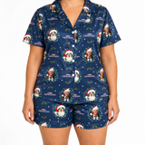 A model wearing a matching navy blue pajama set with a Christmas-themed print featuring Santa Claus, dogs, holly leaves, and the text 'CHRISTMAS WALKER'.
