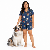 A woman wearing a navy blue pajama set with a Santa Claus and dog print stands next to a fluffy Australian Shepherd dog on a white studio background.