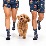 A dog walking between two cropped human legs wearing matching navy Christmas-themed socks and shorts with dog-face prints and Santa hats, on a clean white studio background.