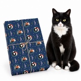 A black and white cat sits beside a wrapped gift box on a clean white background. The gift box is wrapped in navy blue Christmas-themed wrapping paper with a Santa Claus and penguin pattern, tied with twine.