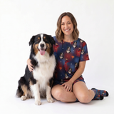 A woman and her Australian Shepherd dog sit together on a white studio floor, wearing matching Supergirl-themed pajamas with dog-print graphics.