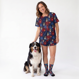 A woman wearing a matching pajama set with a supergirl logo and dog print stands next to a black, white, and tan dog on a white studio background. She is also wearing matching patterned socks.