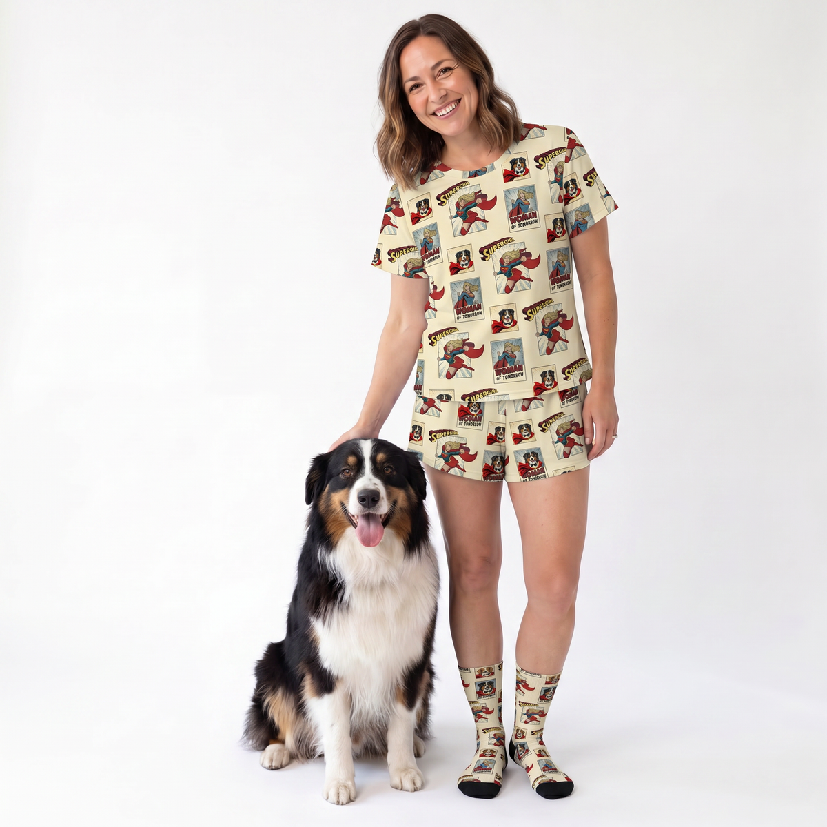 A woman wearing a matching beige pajama set with a comic book style print stands next to a black, white, and tan dog on a white studio background. The set includes a short-sleeve top and shorts, paired with matching socks.
