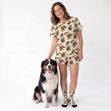 A woman wearing a matching beige pajama set with a comic book style print stands next to a black, white, and tan dog on a white studio background. The set includes a short-sleeve top and shorts, paired with matching socks.