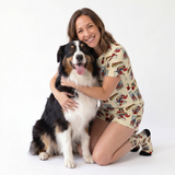 A woman kneeling on a white studio floor hugging a black, white, and tan dog. She is wearing a short-sleeve pajama romper and matching socks with a vintage comic book cover print featuring Supergirl.