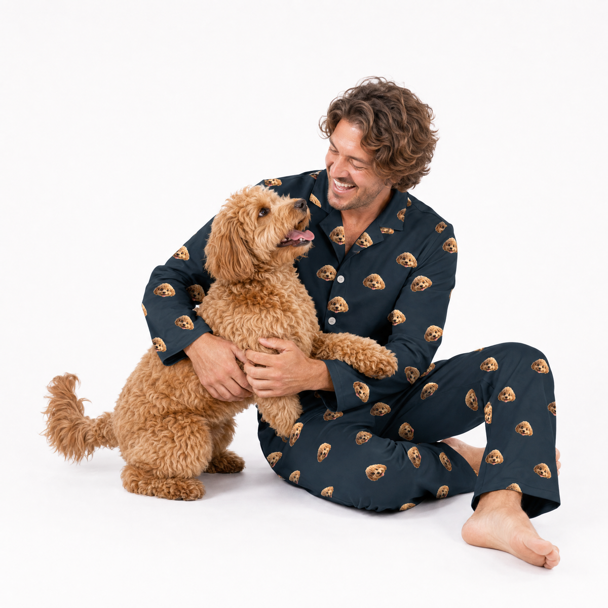 Man in black pajamas with dog pattern sitting on a white background, holding a brown dog.