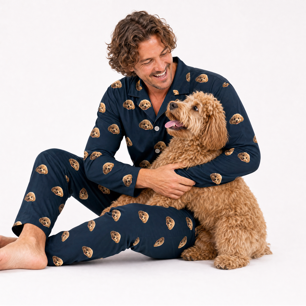 Man wearing black pajamas with dog patterns, sitting with a brown dog on a white background
