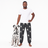 Man wearing black plaid pajama pants with white dog pattern, standing next to a Dalmatian dog on a white background.