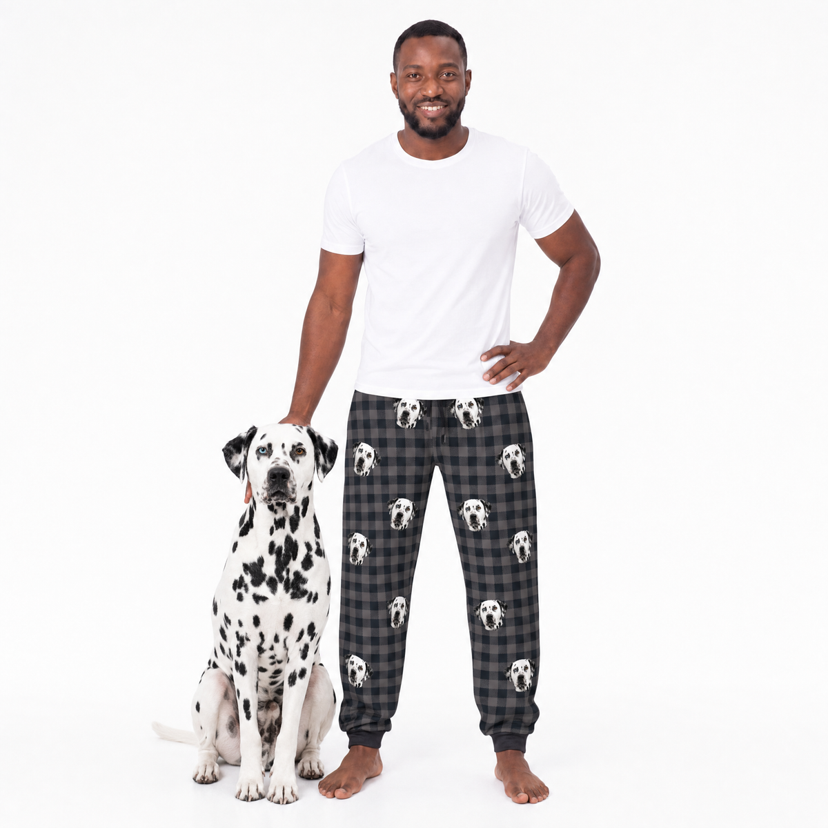 Man wearing black plaid pajama pants with white dog pattern, standing next to a Dalmatian dog on a white background.