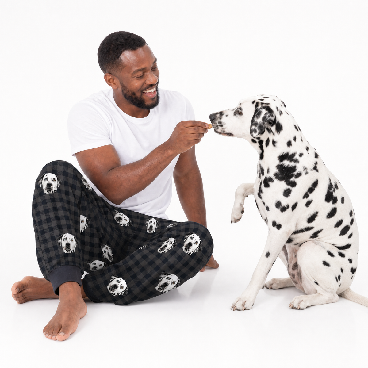 Man in pajama pants with dog pattern feeding a Dalmatian dog on a white background