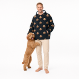 Man wearing a black blanket hoodie with dog face patterns, standing next to a brown dog on a white background