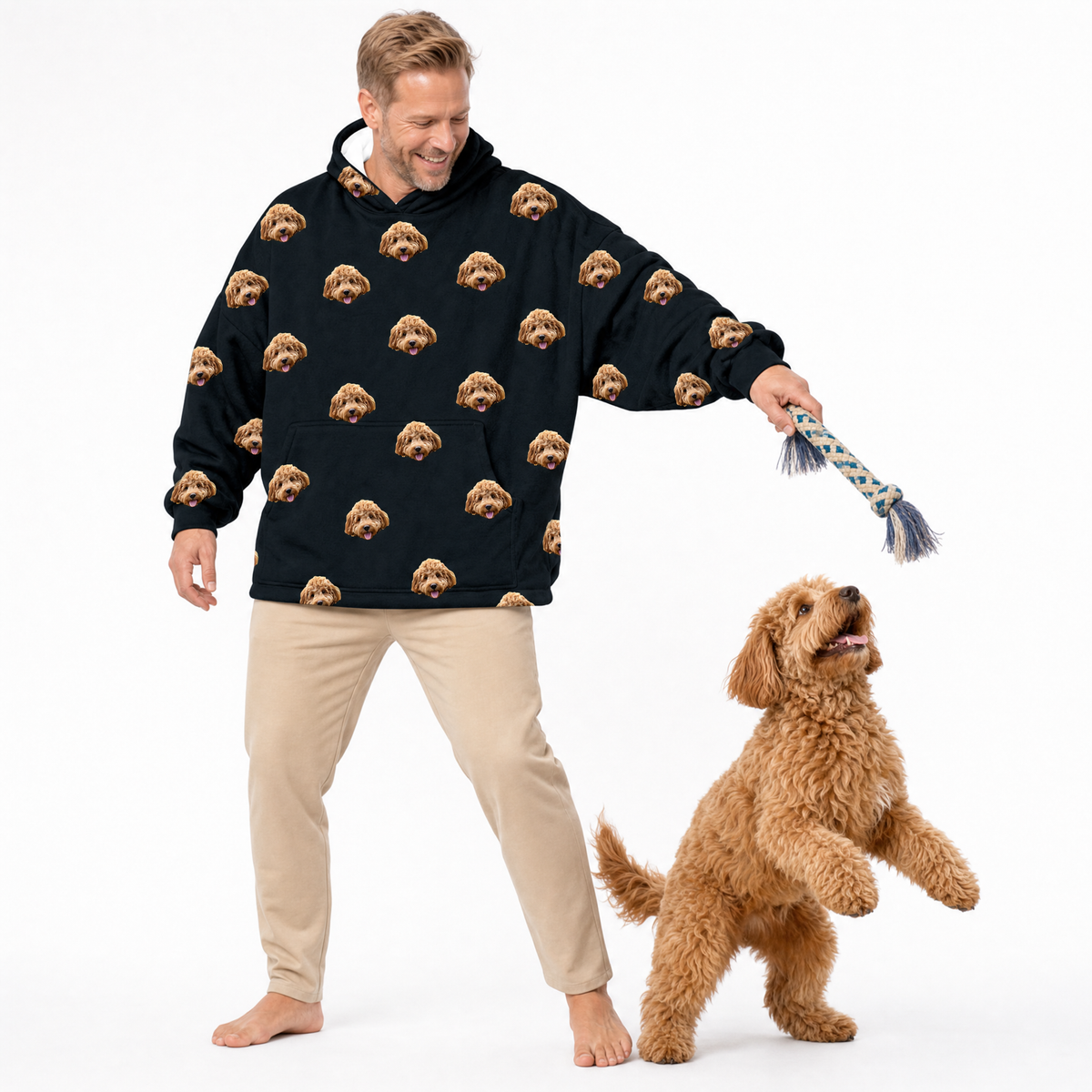 Man wearing a black blanket hoodie with dog pattern and beige pants, standing next to a brown dog on a white background
