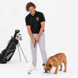 A man in a black polo shirt and light gray trousers holds a golf club next to a black golf bag, with a brown bulldog sniffing a golf ball on the ground, set against a solid white studio background.