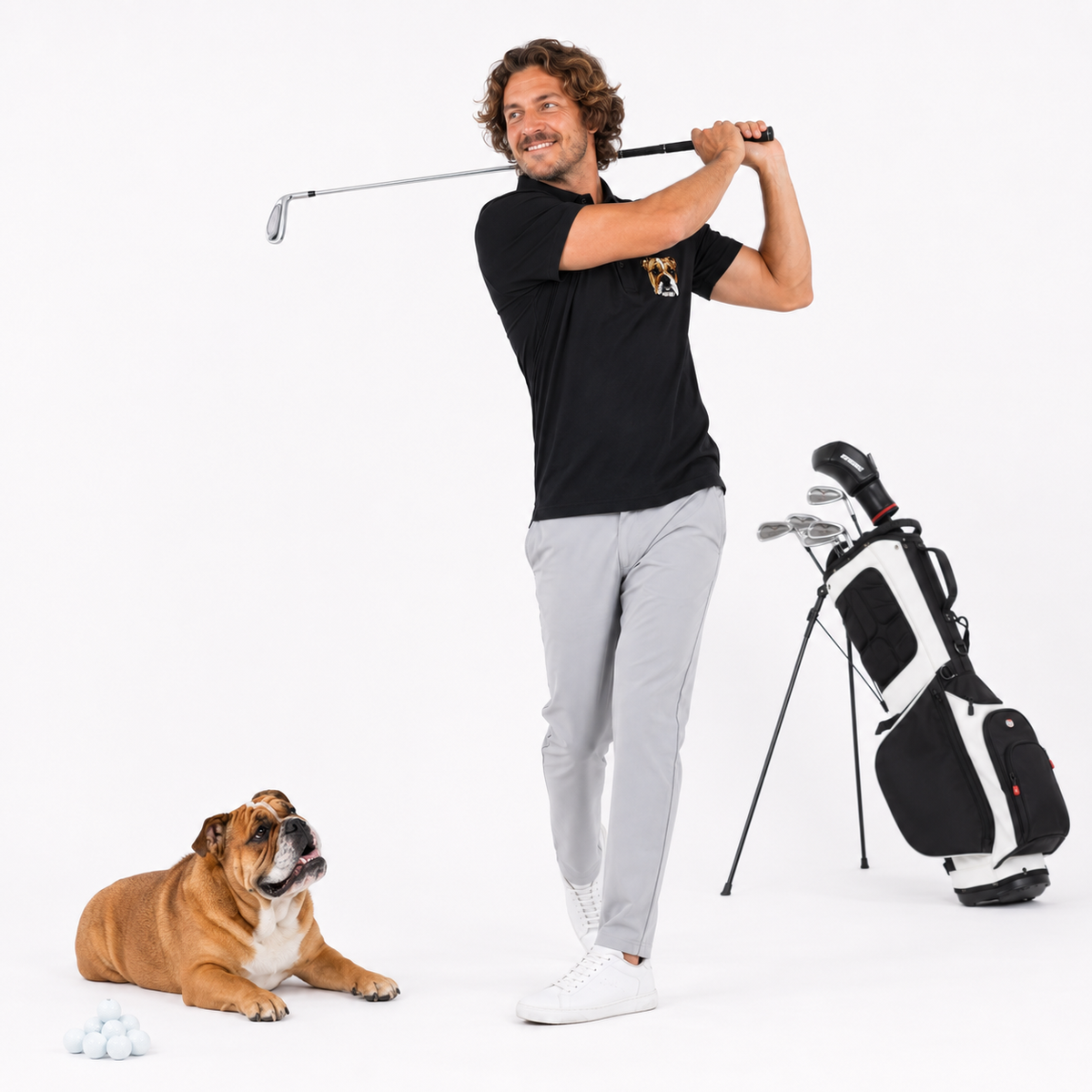 A man in a black polo shirt and light gray trousers is captured mid-swing with a golf club, standing next to a black and white golf bag on a white studio background. A brown bulldog lies on the ground in the foreground next to golf balls.