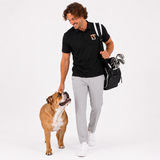 A man wearing a black polo shirt, light gray trousers, and white sneakers walks while petting a brown and white dog. He is carrying a black golf bag with clubs over his shoulder on a clean white studio background.
