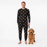 Man wearing a black jogger pajama set with a dog pattern, standing next to a brown dog on a white background.