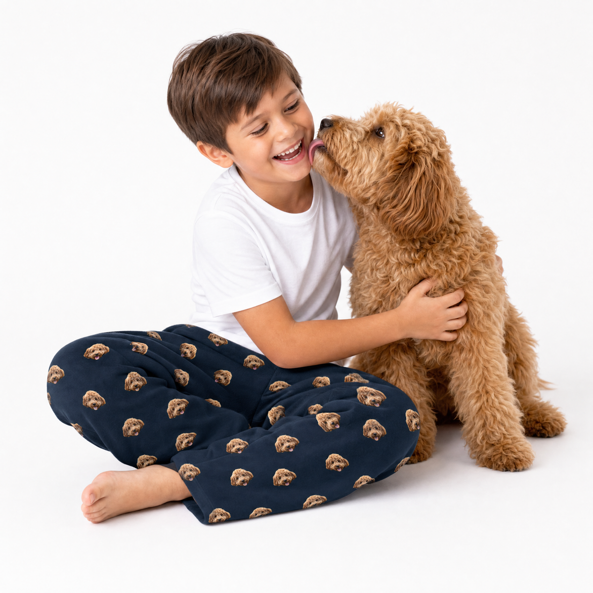 Child wearing black pajama pants with dog patterns, sitting on a white background with a brown dog.