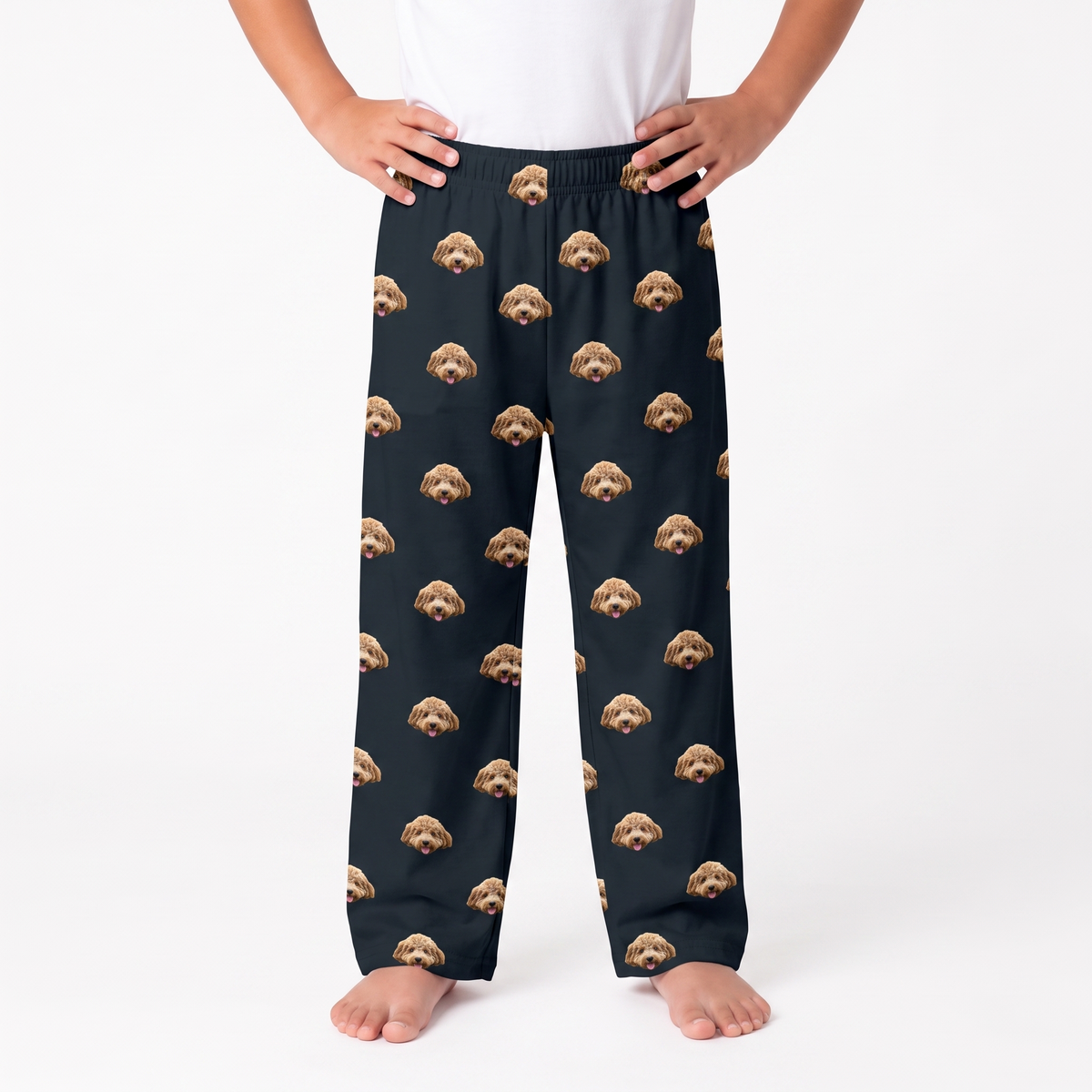 A person wearing black pajama pants with an all-over repeating print of small tan and brown dog faces, standing barefoot against a clean white studio background.