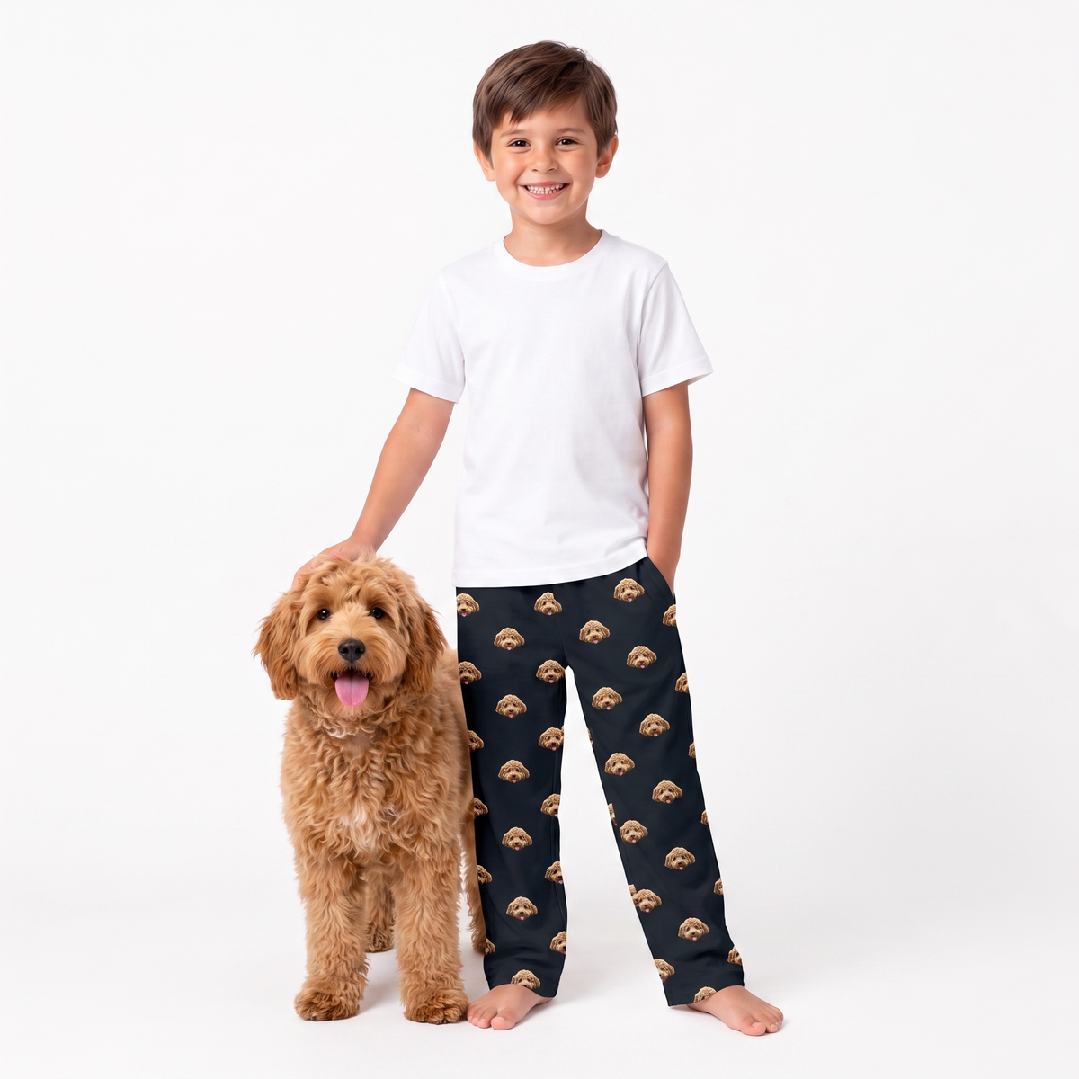 A young boy stands barefoot next to a fluffy dog, wearing a white t-shirt and black pajama pants with a repeating pattern of small tan and brown dog faces. The background is a clean white studio backdrop.