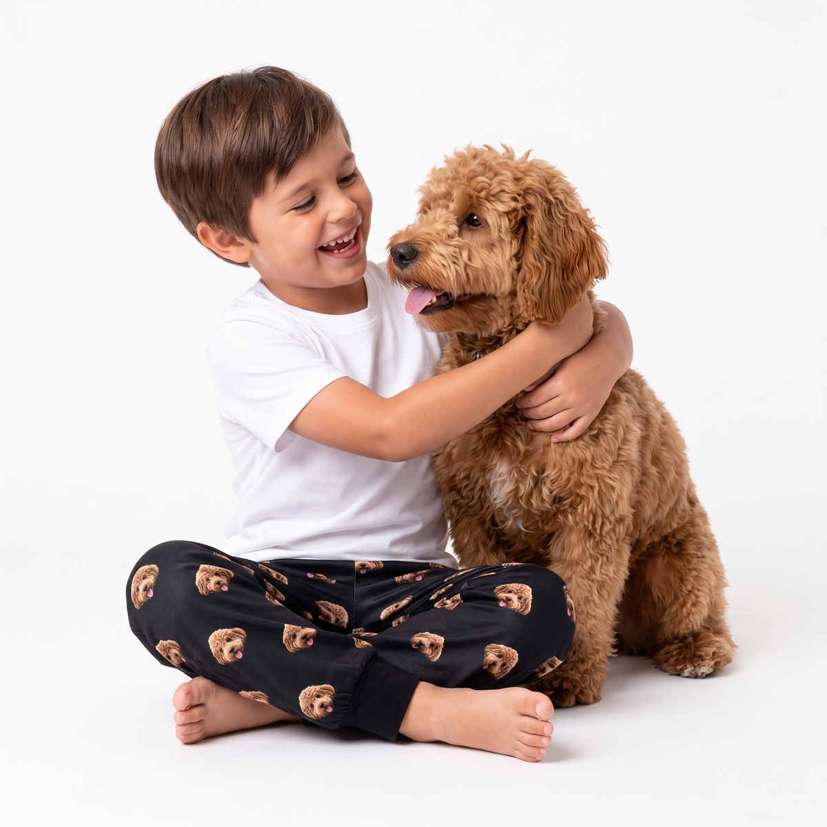 A young boy with brown hair sits cross-legged on a white studio floor, hugging a fluffy light brown dog. The boy is wearing a white short-sleeve t-shirt and black pajama pants with a repeating pattern of small dog faces.