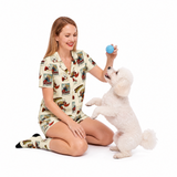A woman and her small white dog are sitting on a white studio background. The woman is wearing a matching pajama set with a comic book vintage-style illustrated print, and she is holding a blue ball for the dog.
