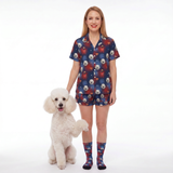 A woman stands next to a small white poodle wearing a matching pajama set with a Supergirl-theme and dog print, including a short-sleeve button-up shirt and shorts, along with matching socks, against a plain white studio background.